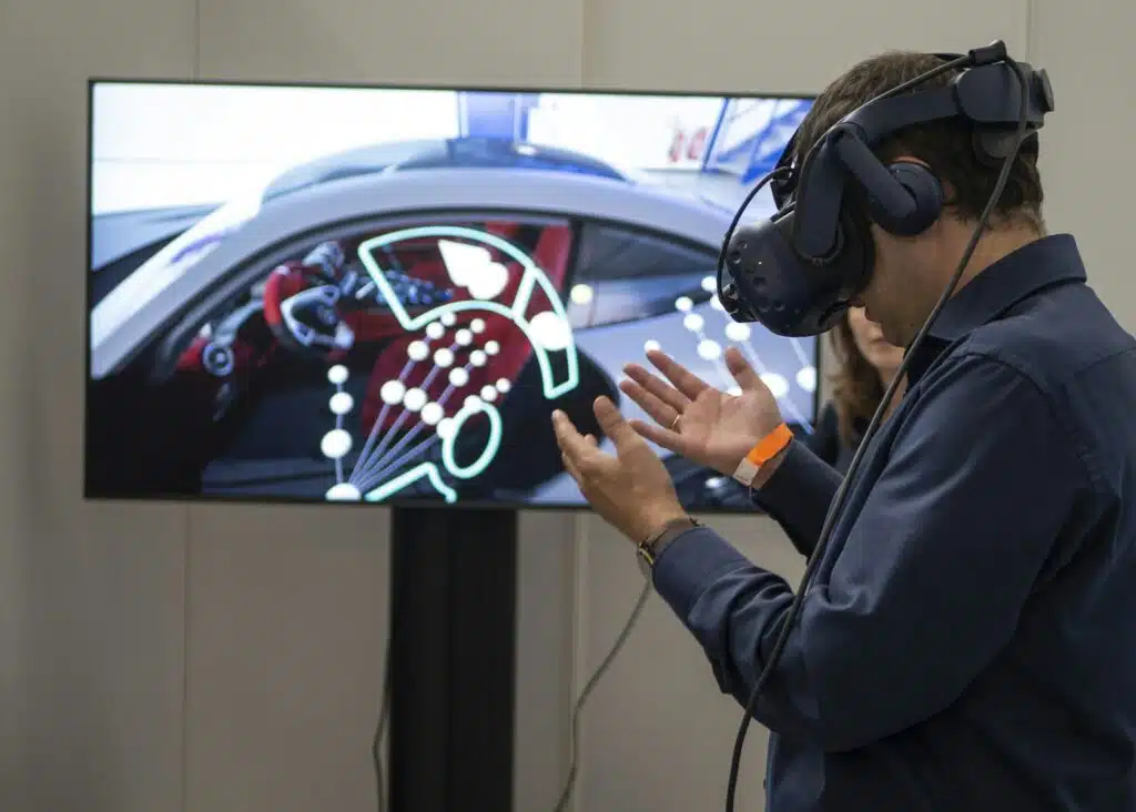 Homme utilisant un casque de réalité virtuelle HTC Vive interagissant avec l'interface futuriste d'une voiture affichée sur un grand écran.